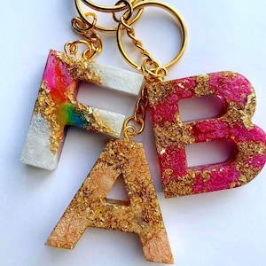 May include: A gold keychain with three resin letters that spell "FAB". The letters are filled with gold flakes and have different coloured backgrounds: white, pink, and peach.
