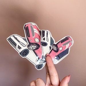 Peut inclure: Plusieurs autocollants en forme de voiture, roses et blancs, avec des détails noirs et gris. Les autocollants présentent différents modèles de voitures, dont un cabriolet classique et une voiture de sport moderne. Les autocollants sont tenus par les doigts d'une personne.
