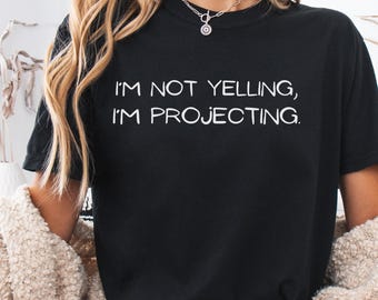Puede incluir: Camiseta negra con el texto blanco "I'M NOT YELLING, I'M PROJECTING." La camiseta tiene cuello redondo y mangas cortas. La modelo lleva un collar plateado y una rebeca color crema.