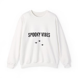 Peut inclure: Un sweat-shirt blanc à col rond avec l'inscription "SPOOKY VIBES" en noir, dans une police dégoulinante. En dessous, trois petites araignées noires suspendues à des fils, sur le thème d'Halloween.
