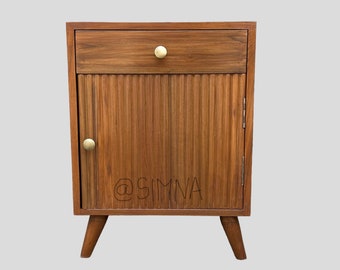 Puede incluir: Una mesita de noche de madera con un cajón y una puerta de armario con ranuras verticales. La mesita de noche tiene pomos de latón y patas cónicas. La puerta del armario tiene el texto "@SIMMA" escrito en ella.