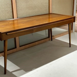 May include: A wooden desk with two drawers and a light brown finish. The desk has a simple design with straight lines and a rectangular shape. The desk is sitting on a white floor.