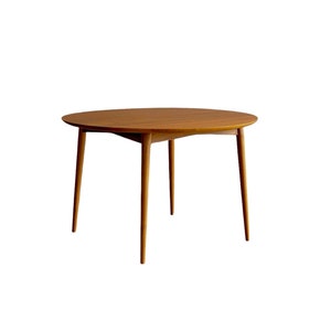 Puede incluir: Una mesa redonda de madera con acabado marrón claro. La mesa tiene una superficie lisa y cuatro patas anguladas. La mesa tiene aproximadamente 100 cm de diámetro, adecuada para un comedor o sala de estar.