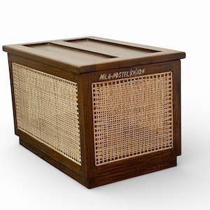 May include: A rectangular wooden storage chest with a woven cane panel on the front and sides. The top is flat with two recessed lines. The chest is stained a dark brown color. The text "MLA-HOSTEL(CH)124" is written on the side.