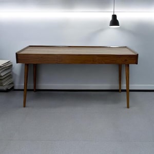 May include: A wooden desk with three drawers and tapered legs. The desk is made of dark wood and has a natural finish.