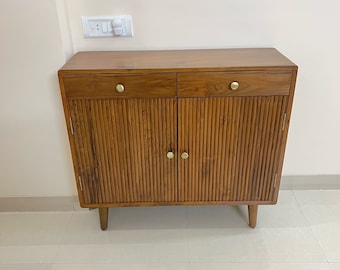 May include: A wooden cabinet with two doors and two drawers. The cabinet has a light brown finish and gold-colored hardware. The cabinet is sitting on a white tiled floor.