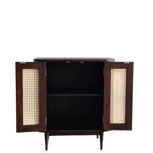 May include: A dark brown wooden cabinet with two doors and a shelf inside. The doors are made of woven cane and are open, revealing the empty shelf. The cabinet has a simple design with tapered legs.