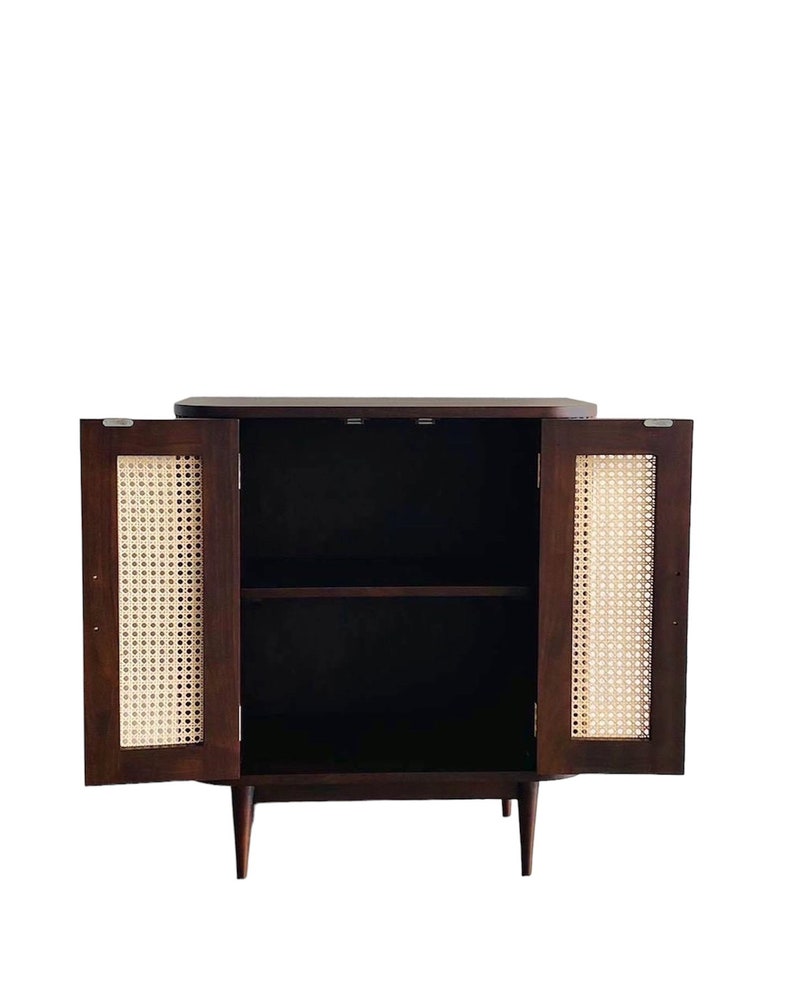 May include: A dark brown wooden cabinet with two doors and a shelf inside. The doors are made of woven cane and are open, revealing the empty shelf. The cabinet has a simple design with tapered legs.