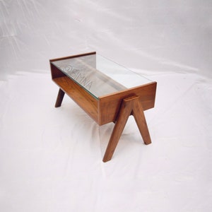 Puede incluir: Una mesa de centro de madera con una tapa de vidrio y un estante debajo. La mesa tiene un diseño único en forma de A y está hecha de madera oscura.