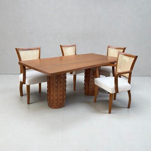 Puede incluir: Un juego de comedor moderno de mediados de siglo. La mesa rectangular de madera tiene dos pedestales cilíndricos texturizados. Cuatro sillas con respaldos de mimbre y asientos tapizados blancos rodean la mesa. La madera tiene un acabado cálido y natural.