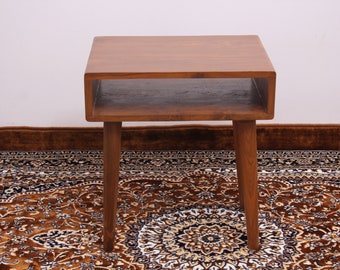 Puede incluir: Una mesa auxiliar cuadrada de madera con un estante. La mesa tiene un acabado de madera natural y está sobre una alfombra estampada.