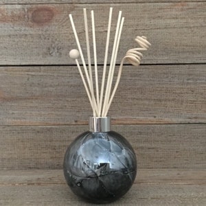 May include: A decorative reed diffuser with a spherical, marbled grey glass bottle and a silver-coloured neck. The diffuser features several light-coloured reeds, a wooden ball, and a decorative swirl. The background is a wooden surface.