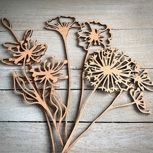 May include: A set of five laser-cut wooden flower shapes, including a dandelion, two poppies, and two smaller flowers. The flowers are all in a natural wood colour and have intricate details.