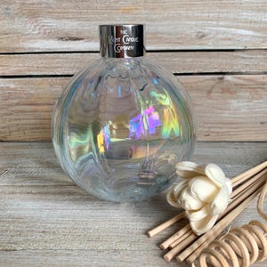 May include: A clear glass diffuser bottle with a silver cap that says "The Kent Candle Company". The bottle has a textured, iridescent surface and is sitting on a wooden surface. There are wooden diffuser sticks and a white flower in the background.