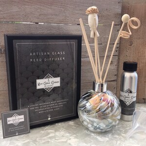 May include: Artisan glass reed diffuser with a silver and white swirl design. The diffuser is packaged in a black box with the text "Artisan Glass Reed Diffuser" and "Kent Candle Company" printed on it. The diffuser comes with a set of reeds and a bottle of fragrance oil.