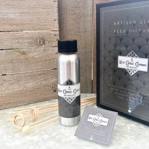 May include: A silver metal bottle with a black lid and a label that reads "The Kent Candle Company Pure Indulgence Oak & Leather 100 ml Diffuser Refill". The bottle is sitting on a white surface with a wooden stick and a small card that reads "The Kent Candle Company Pure Indulgence Extraordinary is our ordinary!"