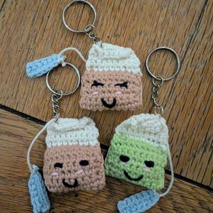 May include: Three crochet tea bag keyrings. Each keyring features a smiling tea bag design with a light blue tag and a silver ring. The tea bags are in shades of brown, green, and white. The keyrings are displayed on a wooden surface.