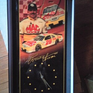 May include: A black and gold clock with a racing theme. The clock features a picture of a race car driver, a race car, and the driver's signature, "Ernie Irvan".