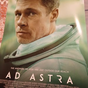 以下が含まれることがあります： ブラッド・ピット主演の映画「アド・アストラ」のポスター。ポスターには、宇宙服を着たブラッド・ピットが横を向いて写っている。映画のタイトルは、ポスターの下部に大きな白い文字で書かれている。映画のキャッチフレーズは「私たちが求める答えは、手の届かないところにあります。」