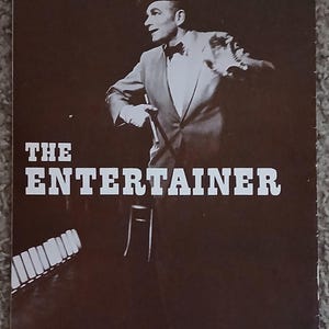 Può includere: Una fotografia in bianco e nero di un uomo in abito e cappello che tiene un bastone. Il testo "THE ENTERTAINER" è stampato in lettere bianche su uno sfondo marrone.