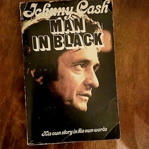 May include: Vintage paperback book titled "Johnny Cash Man in Black" featuring a portrait of Johnny Cash on the cover. The book's title is in bold white letters, with the subtitle "His own story in his own words" below the portrait.