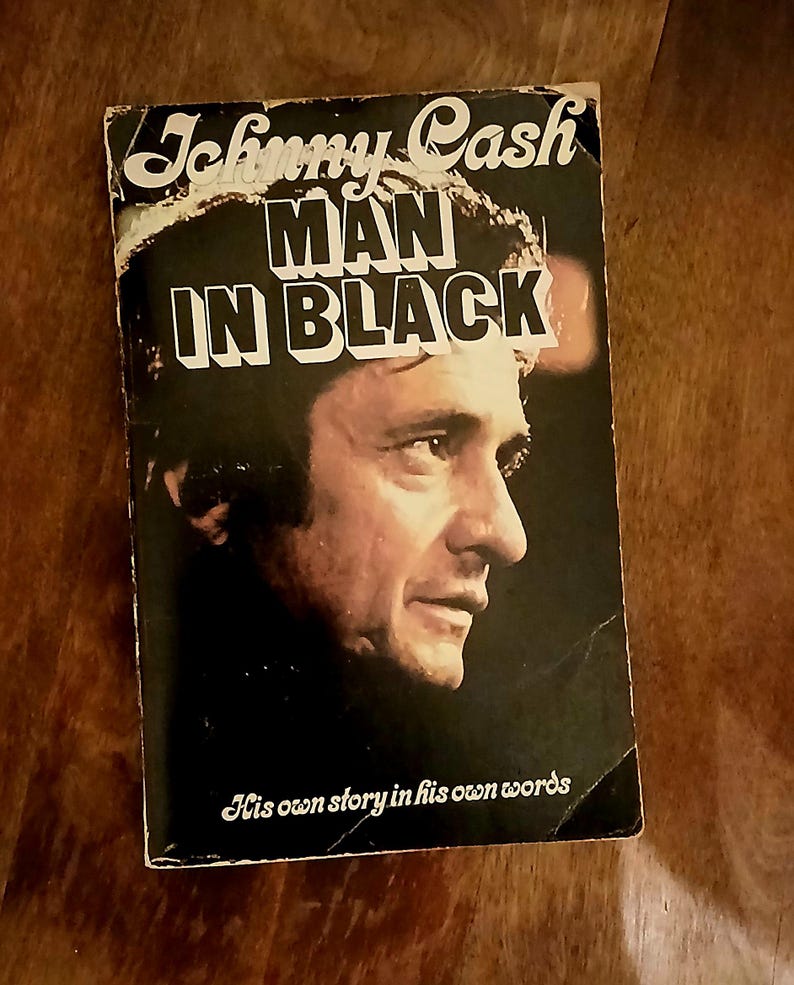 May include: Vintage paperback book titled "Johnny Cash Man in Black" featuring a portrait of Johnny Cash on the cover. The book's title is in bold white letters, with the subtitle "His own story in his own words" below the portrait.