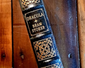 Puede incluir: Un libro de tapa dura vintage titulado "DRACULA" de Bram Stoker. El libro tiene una cubierta azul oscuro con detalles y texto decorativos dorados. El lomo presenta diseños dorados ornamentados.