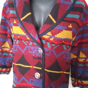 Op de afbeelding: Een rood, zwart, geel, blauw en groen gepatroneerd jack met een shawlkraag en twee zilveren knopen. Het jack is gemaakt van geweven stof.