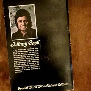 May include: A vintage book cover featuring a portrait of Johnny Cash in a black and white frame. The title "Johnny Cash" is displayed in a stylized font, with text describing his impact on American culture. The cover is black with white text.
