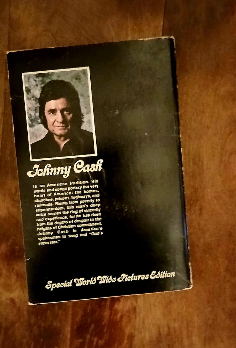 May include: A vintage book cover featuring a portrait of Johnny Cash in a black and white frame. The title "Johnny Cash" is displayed in a stylized font, with text describing his impact on American culture. The cover is black with white text.