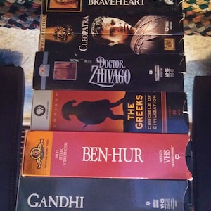 Może przedstawiać: Stos kaset VHS z tytułami filmów, takimi jak "Braveheart", "Kleopatra", "Doktor Żywago", "The Greeks", "Ben-Hur" i "Gandhi". Kasety mają różne kolory i wzory, z tekstem i obrazami na okładkach.