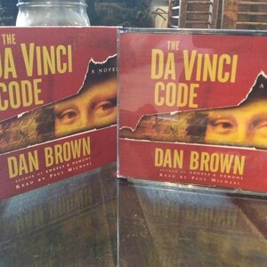 May include: Two red audio books with the title "The Da Vinci Code" by Dan Brown. The cover features a close-up of the Mona Lisa's eyes.