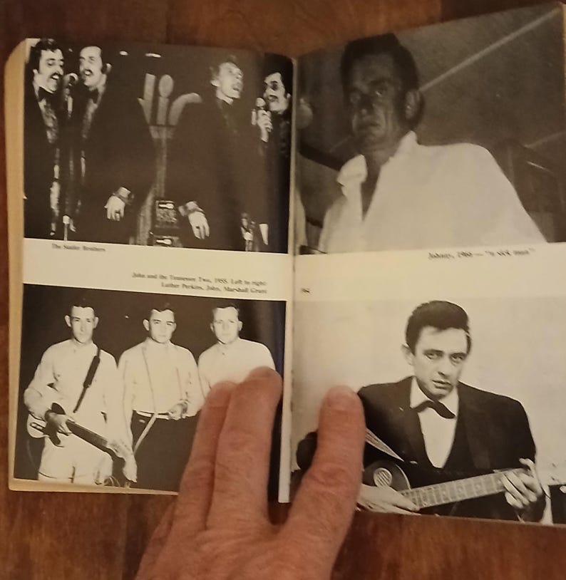 May include: A black and white open book featuring vintage photographs of musicians. The images include The Statler Brothers, Johnny Cash, and John and the Tennessee Two. The book is open, with a hand partially visible.