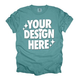 May include: A teal blue t-shirt with the text "YOUR DESIGN HERE" in white. The text is stylized with a heart and stars.