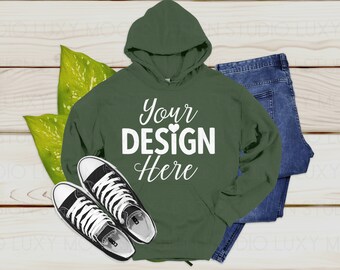 May include: A dark green hooded sweatshirt with the text "Your Design Here" in white. The sweatshirt is laying on a wooden surface with a pair of black canvas shoes and blue jeans.