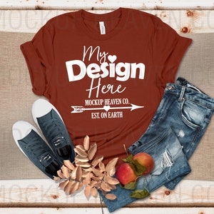 May include: A rust-colored t-shirt with the text "My Design Here" and "Mockup Heaven Co. Est. On Earth" in white. The t-shirt is laying on a wooden surface with a pair of blue jeans, two dark blue sneakers, and a sprig of gold leaves and red apples.