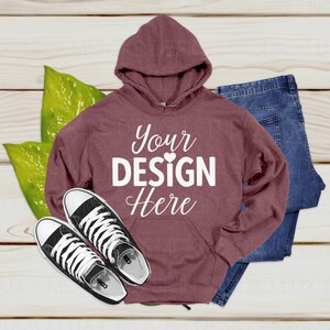 Op de afbeelding: Een maroon hoodie met een grote witte afbeelding die "Your Design Here" op de voorkant zegt. De hoodie ligt op een houten oppervlak met een paar zwarte sneakers en een blauwe jeans.