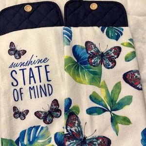 May include: Two white kitchen towels with a navy blue top and wooden button. The towels feature a butterfly and leaf design with the text "sunshine STATE OF MIND" in blue. The butterflies have red, blue, and white details.