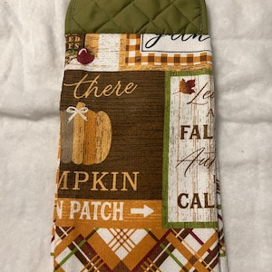 May include: A hanging kitchen towel with a quilted olive green top and a wooden button closure. The towel features a fall-themed design with pumpkins, leaves, and the words "Pumpkin Patch" and "Fall". The bottom is white.