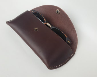 May include: A brown leather sunglasses case with a flap closure and a snap button. The case is open and a pair of tortoiseshell sunglasses are inside.