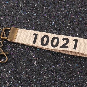 May include: A beige fabric keychain with a metal ring and clasp. The keychain has the number "10021" printed in black.