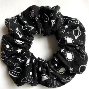 May include: Black scrunchie with a white space-themed print featuring stars, planets, rockets, and UFOs.