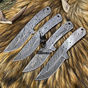 Peut inclure: Quatre lames de couteau en acier Damas avec une finition martelée. Les lames sont disposées en rangée, les pointes pointant vers la droite. Les lames sont sur un fond de fourrure brun et blanc. Le texte "Vikings Norseman" est visible sur les lames.