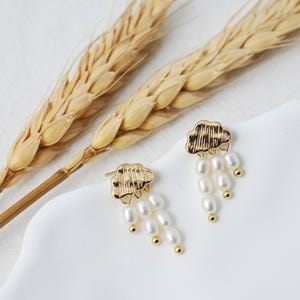 Può includere: Un paio di orecchini dorati con un design a nuvola e gocce di perle. Gli orecchini sono su una superficie bianca con steli di grano.