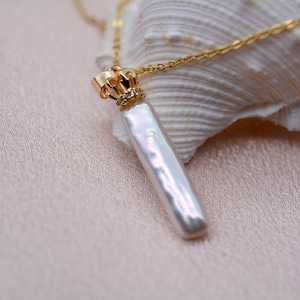 Peut inclure: Un collier en chaîne dorée avec un pendentif en forme de couronne et une longue perle baroque blanche. La perle est attachée à la couronne par un connecteur en or.