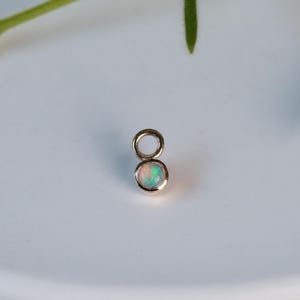 Può includere: Un piccolo ciondolo dorato con una gemma di opale rotonda e iridescente. L'opale mostra un mix di tonalità rosa pallido, verdi e blu. Il ciondolo ha un piccolo anello nella parte superiore per il fissaggio a una catena o un braccialetto. Il gioiello è posto su uno sfondo bianco.