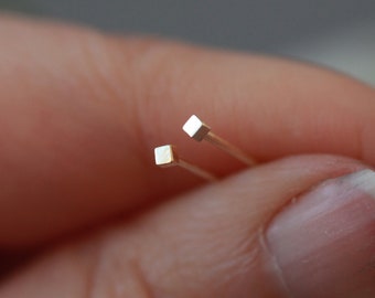 Pode incluir: Um par de brincos minimalistas quadrados de ouro. Os brincos são pequenos e delicados, com uma forma quadrada simples.