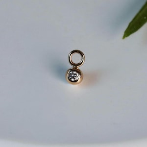May include: A small, gold-colored pendant with a round, bezel-set diamond. The pendant has a loop at the top for attaching to a chain. The diamond sparkles, and the gold has a warm tone. The pendant is likely for jewelry.