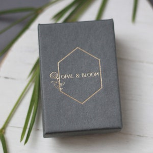 Może przedstawiać: Szkatułka na biżuterię w kolorze szarym z złotym sześciokątnym logo z napisem "OPAL & BLOOM" i stylizowanym wzorem kwiatu.
