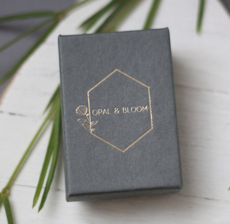 Może przedstawiać: Szkatułka na biżuterię w kolorze szarym z złotym sześciokątnym logo z napisem "OPAL & BLOOM" i stylizowanym wzorem kwiatu.
